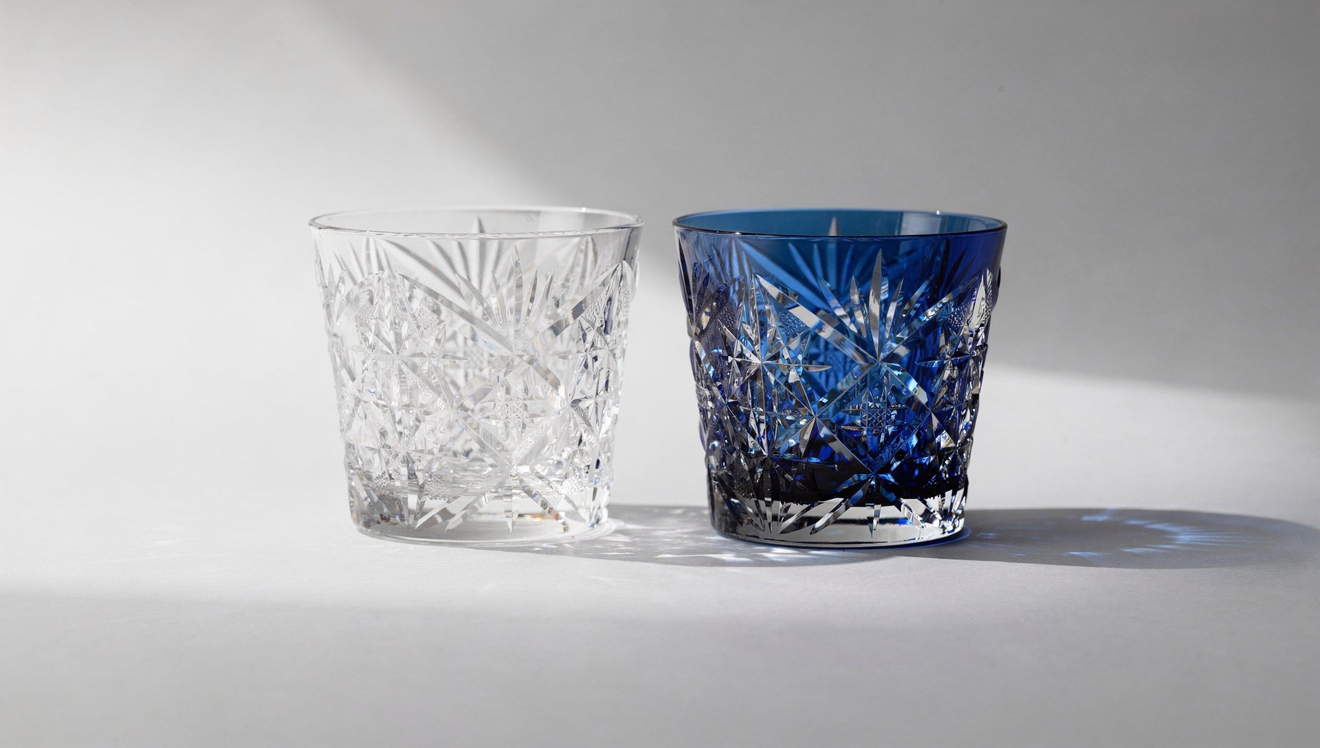 A clear cut-glass rocks glass beside an empty cobalt overlay glass on pale grey.
