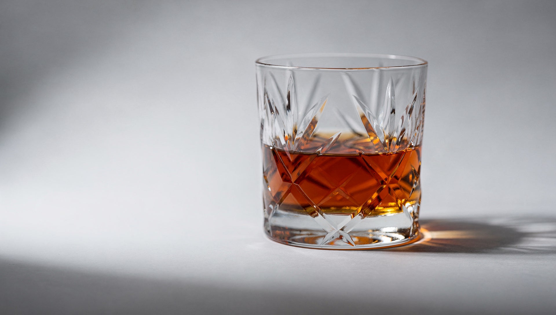 A cut rocks glass with a short neat pour of amber whisky on pale grey.