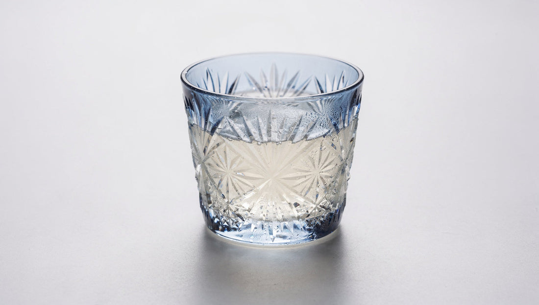 A small Japanese cut-glass sake cup with a chilled pale pour and a hint of condensation on a pale ground.