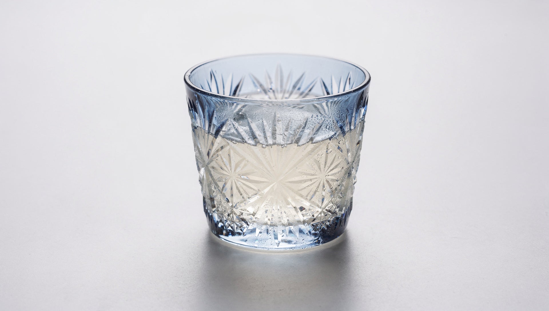 A small Japanese cut-glass sake cup with a chilled pale pour and a hint of condensation on a pale ground.