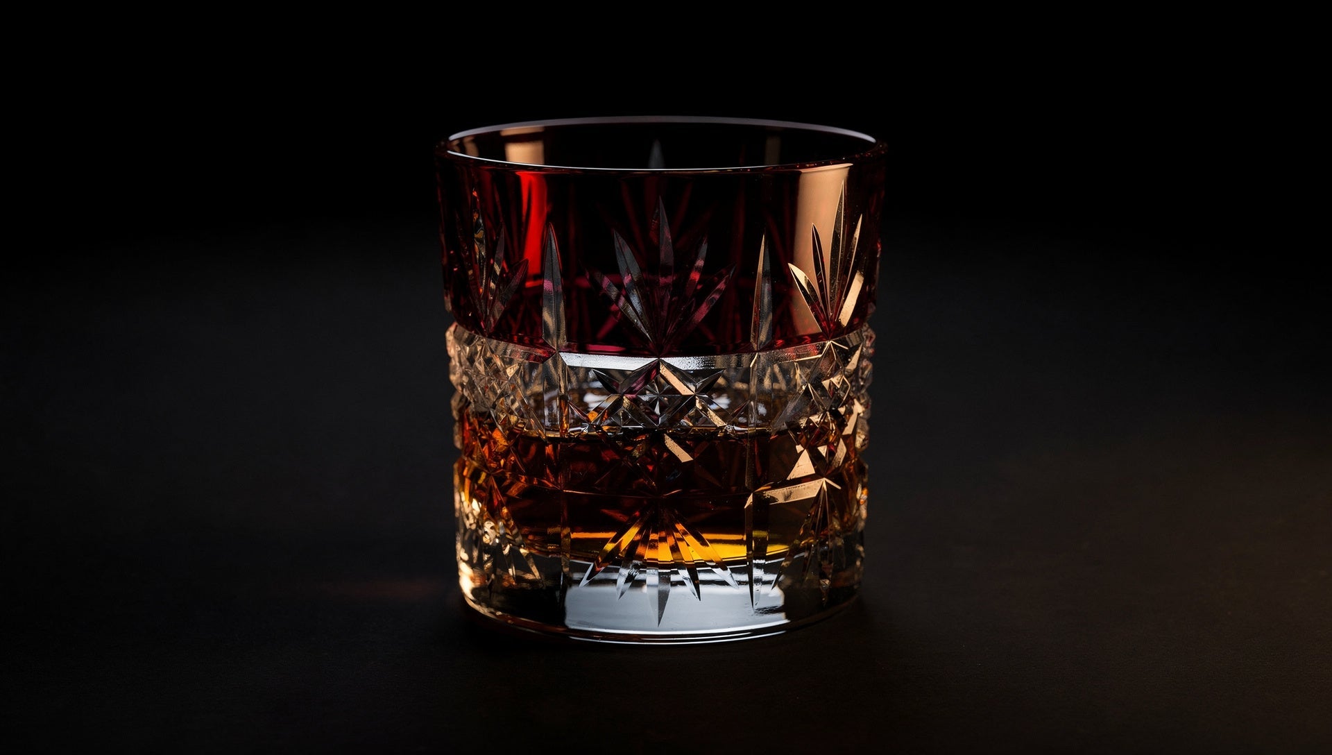 Amber whisky in a ruby or cobalt Japanese cut-glass rocks glass in evening light on an ink ground.