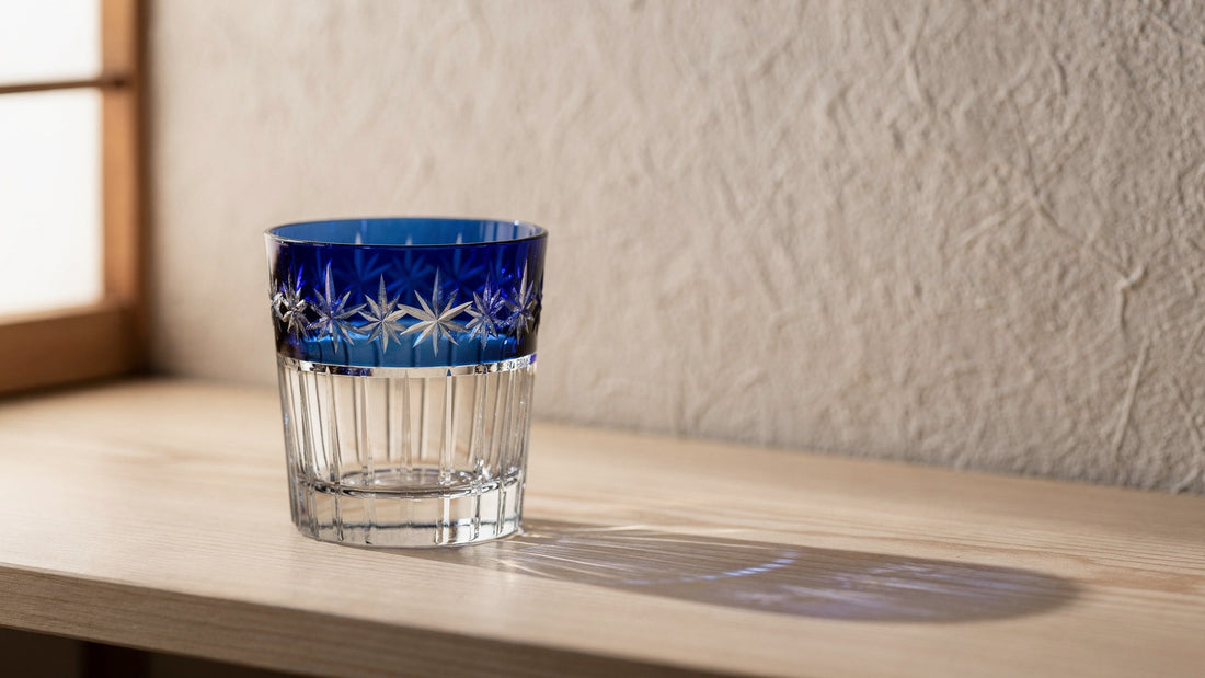An empty cobalt and clear Japanese cut rocks glass on a pale wood sill beside a paper wall, soft window light casting a long blue-tinted shadow.