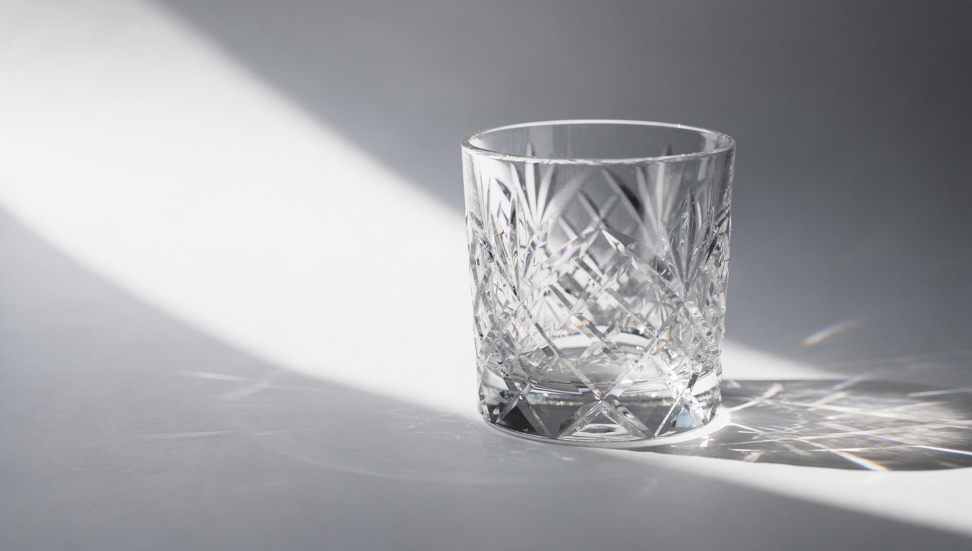A cut-glass tumbler throwing facet light marks and caustics across a pale #f1f1f1 ground.