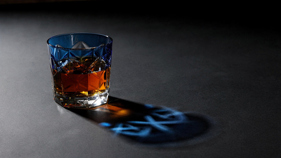 A cobalt Japanese cut rocks glass with a short amber pour on a near-black table under hard caustic light