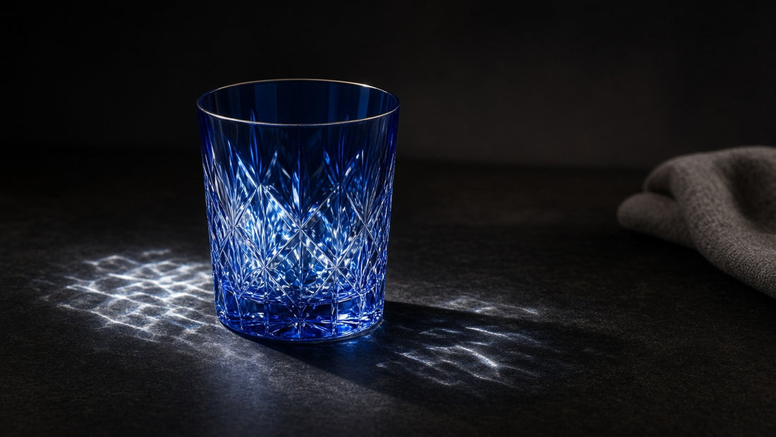 A cobalt Japanese cut rocks glass on a near-black table with hard caustic light across the facets, and a folded cloth at the edge