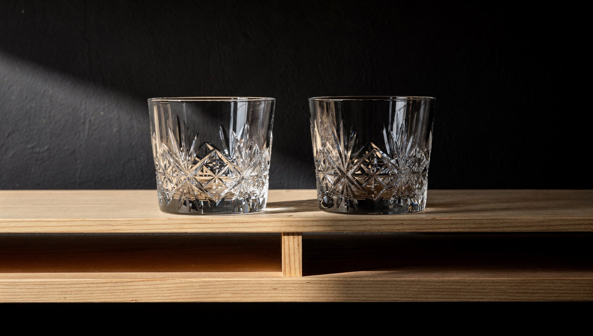 Two Japanese cut-glass rocks glasses stored upright on an open shelf, spaced apart, ink wall and a thin band of daylight.
