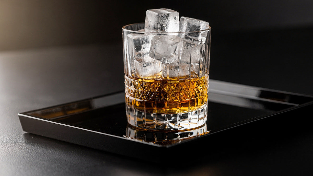 A clear Japanese cut rocks glass packed with clear ice and a pale amber whisky pour on a black lacquer tray, soft side light from the left.