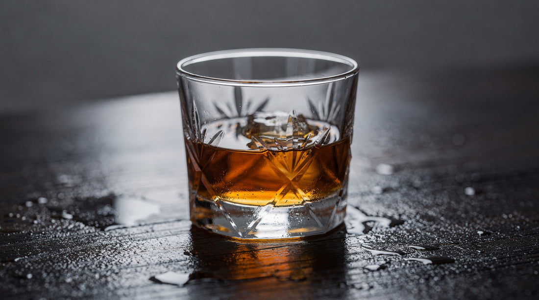 A clear Japanese cut rocks glass with a short amber pour on dark damp wood, cool quiet light, no frost.