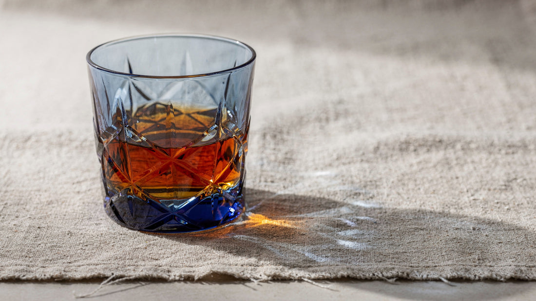 A cobalt Japanese cut rocks glass with a short amber pour on natural linen cloth, soft side light casting caustics on the weave
