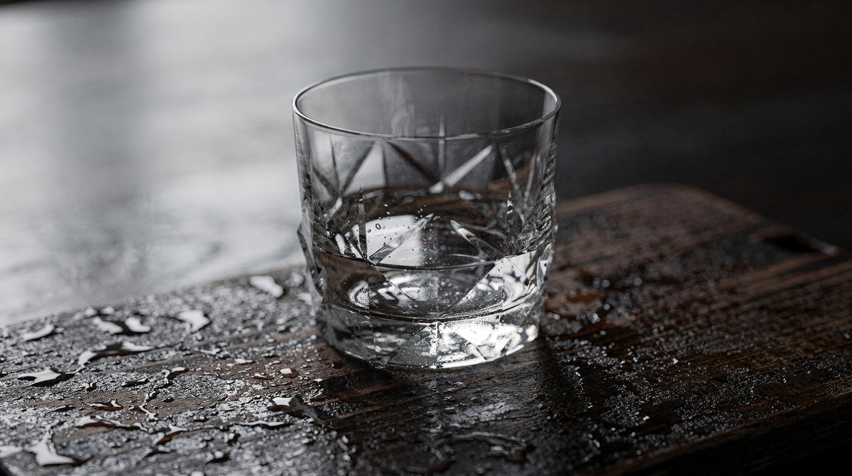 An empty clear Japanese cut rocks glass on dark damp rain-wood, quiet moisture and soft side light.
