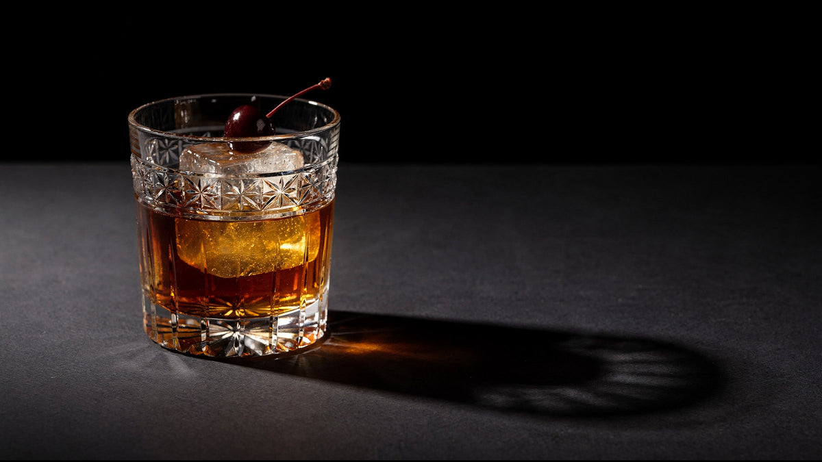 A clear Japanese cut rocks glass with one large ice cube and a short amber pour on a near-black table, hard side light throwing faceted caustics.