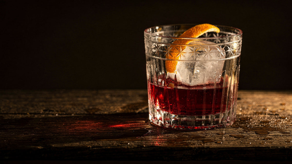 A clear Japanese cut rocks glass with one large ice cube, a deep red pour and a strip of orange peel on dark wet wood, low warm side light casting a red glow
