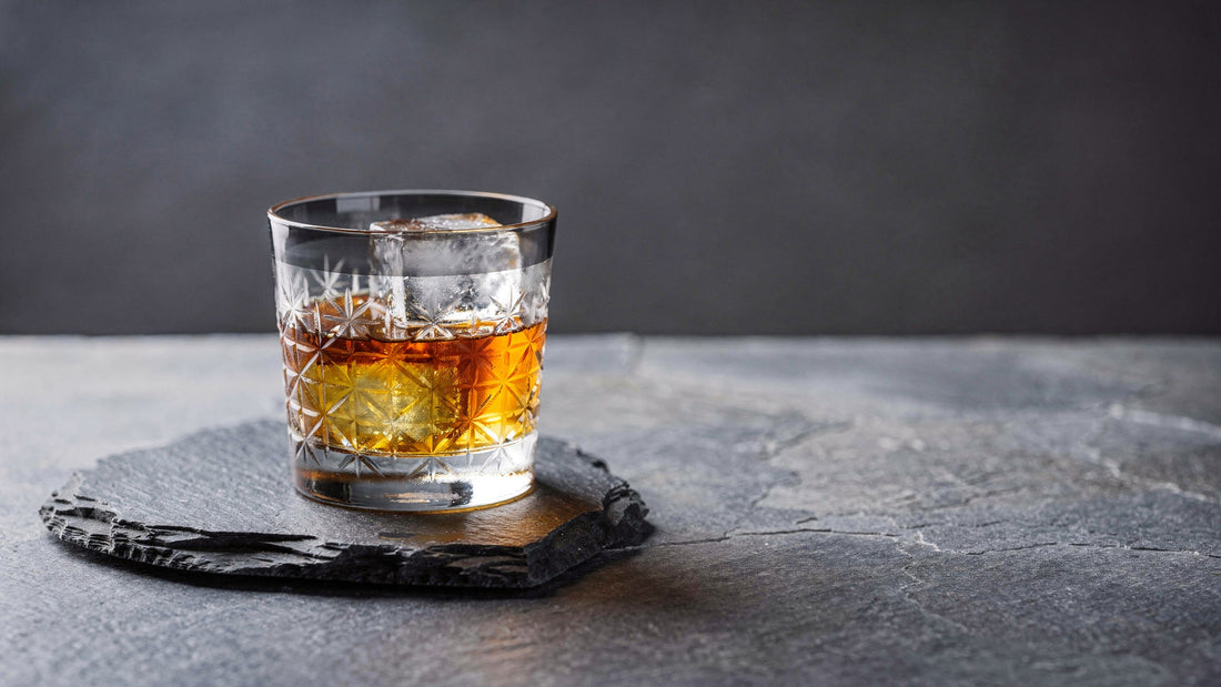 A Japanese cut rocks glass with a smoke-grey band below the rim, one large clear ice cube and an amber cocktail with a darker top layer, on a slate slab under soft side light.