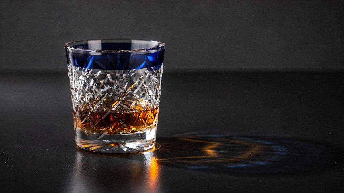A cobalt Japanese cut rocks glass with a short amber pour and no ice on a near-black table, hard side light casting amber and blue caustics.