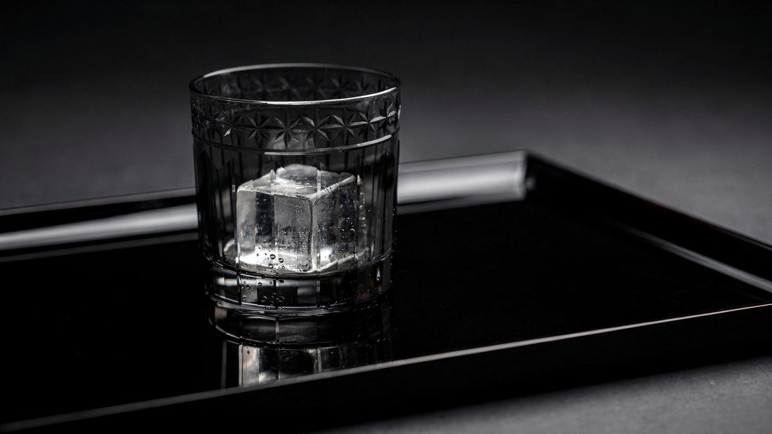A smoke Japanese cut rocks glass with one large clear ice cube and a short clear pour on a black lacquer tray, soft side light and a quiet reflection
