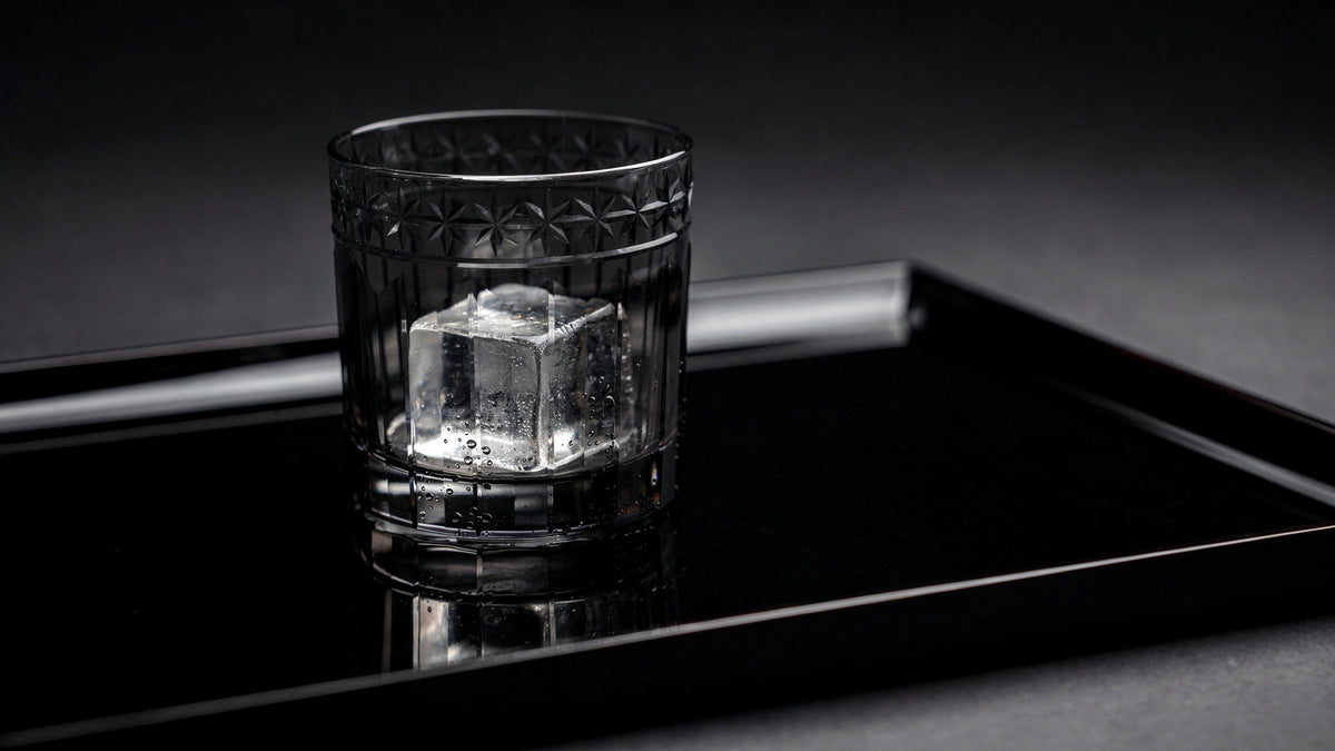 A smoke Japanese cut rocks glass with one large clear ice cube and a short clear pour on a black lacquer tray, soft side light and a quiet reflection