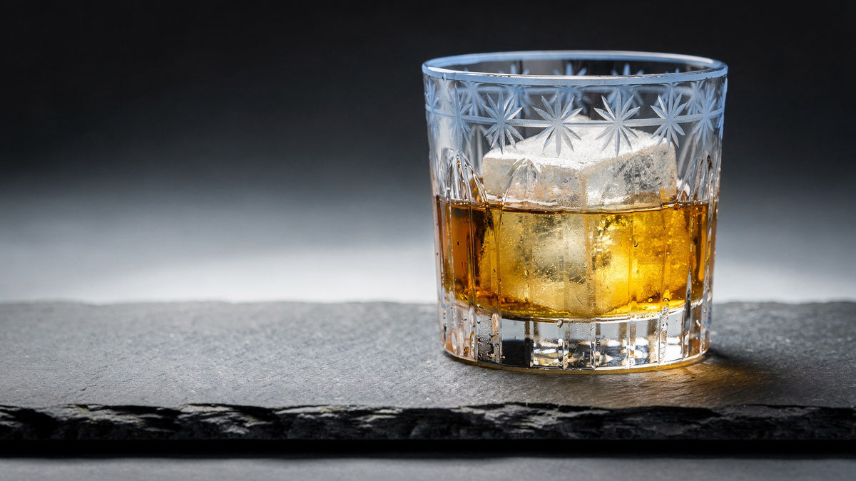 A clear Japanese cut rocks glass with one large ice cube and a short amber pour on dark slate, cool rim light and a warm glow on the stone
