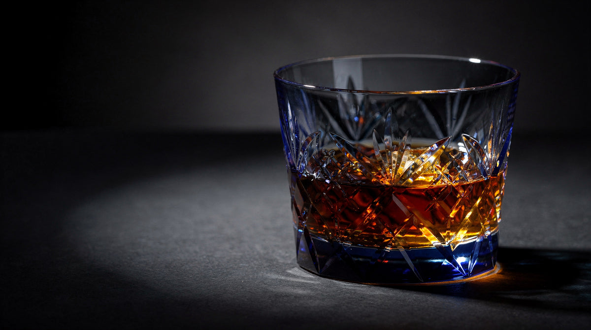 A cobalt Japanese cut rocks glass with a short amber pour on a near-black ink table, hard caustic light on the facets.