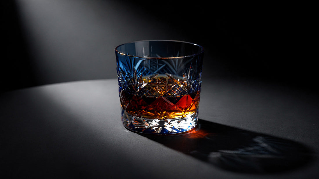 A cobalt Japanese cut rocks glass with a short amber pour and clear headspace on a near-black ink table, hard caustic light on the facets