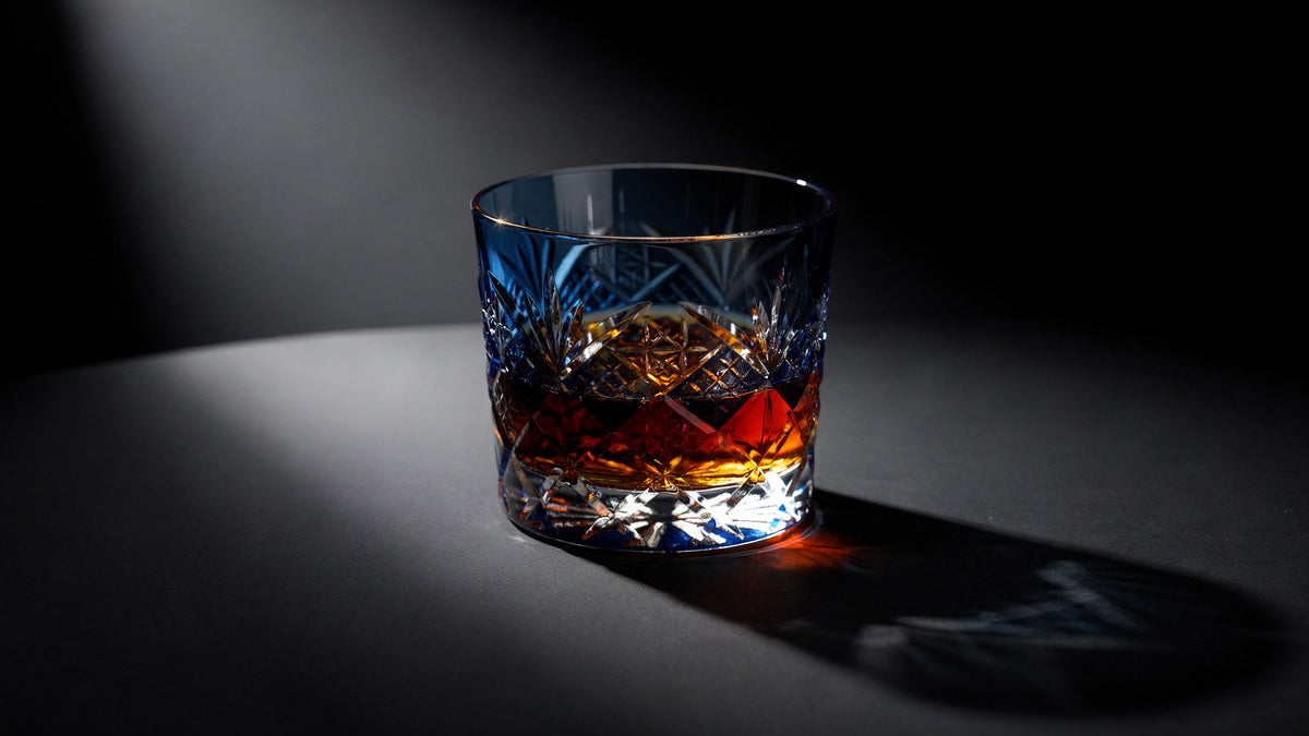 A cobalt Japanese cut rocks glass with a short amber pour and clear headspace on a near-black ink table, hard caustic light on the facets