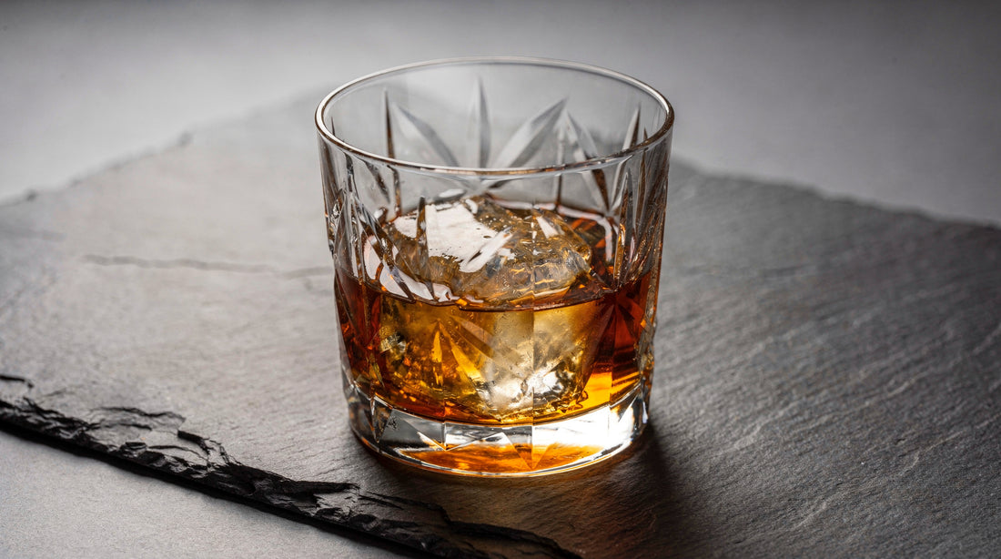 A clear Japanese cut rocks glass with a short amber pour and one large clear ice cube on cool slate, rim light.