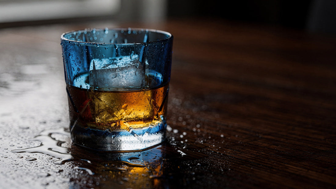 A cobalt Japanese cut rocks glass with a short amber pour and one ice cube on dark damp wood, side light catching condensation on the wall