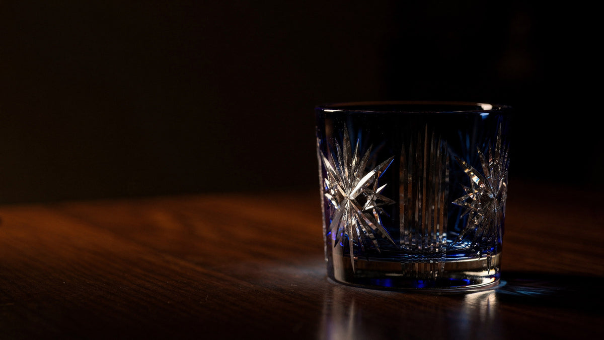 A cobalt Japanese cut rocks glass on a dark wood table in a dim room, a low lamp lighting its facets from the side